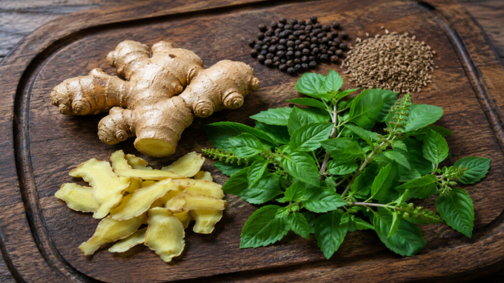 Fresh ginger, tulsi leaves, black pepper, and cumin seeds on a wooden board.